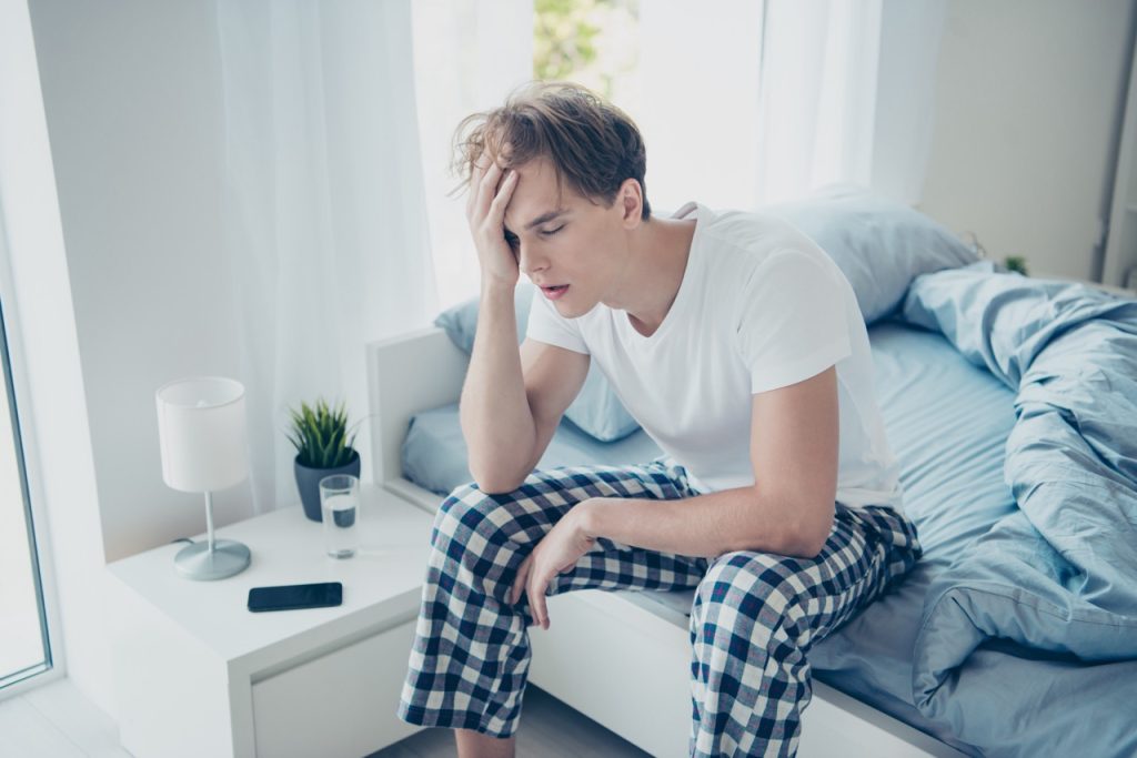 Portrait of exhausted unhappy man after hangover party have terrible fatigue migraine catch cold suffer sit on bed wear checkered plaid pajama in house indoors