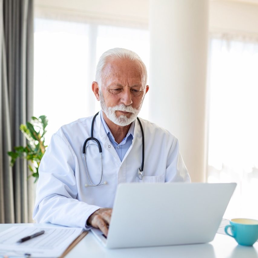 Serious mature doctor using laptop and sitting at desk. Senior professional medic physician wearing white coat and stethoscope working on computer at workplace.